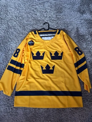  Tre Kronor four nations tröja  - Säljer en gul hockeytröja med Tre Kronor-tryck och nummer 88 på ärmar och rygg. Tröjan har mörkblå detaljer, namntryck 'Nylander' på ryggen och är tillverkad i ett lätt, syntetiskt material. Perfekt för dig som älskar svensk hockey och vill sticka ut på läktaren. Priset är inte fast så hör av dig vid funderingar!