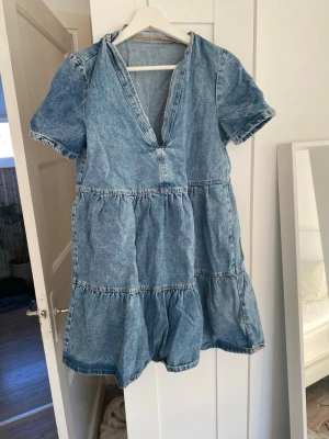 Blå jeansklänning med v-ringning - Snygg blå jeansklänning med korta ärmar och v-ringad hals. Klänningen har volangdetaljer i flera lager och avslappnad passform. Perfekt för dig som gillar en casual och trendig stil. Tillverkad i jeansmaterial som ger en cool och tidlös look.