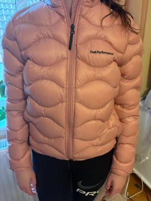 Rosa dunjacka från Peak Performance - Säljer en snygg rosa dunjacka från Peak Performance med vågigt quiltat mönster och hög krage. Jackan har dragkedja framtill och är tillverkad i ett glansigt material som håller dig varm under kalla dagar. Perfekt för dig som vill ha både stil och funktion under våren. Aldrig använts, hängt i garderob sedan köp.  Mycket fint skick.  Storlek: barnstorlek 170 vilket motsvarar en S i vuxenstorlek. En M kan absolut också ha den.  Material: Ankdun/ Polyamid.