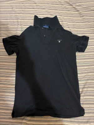 Svart pikétröja från GANT - Klassisk svart pikétröja från GANT med liten logga broderad på bröstet. Tröjan har korta ärmar, krage och knappar vid halsen. Tillverkad i bomull för en skön och luftig känsla. Perfekt till jeans eller shorts.