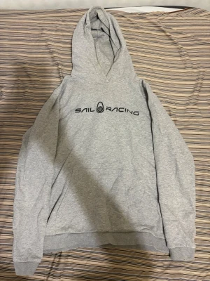 Sail racing hoddie - Snygg grå hoodie från Sail Racing med svart logga tryckt på bröstet. Klassisk modell med huva och känguruficka framtill. Tillverkad i mjukt bomullsmaterial som känns skönt mot huden. Perfekt för en avslappnad och sportig stil.