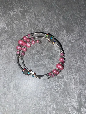 Silvrigt armband med rosa pärlor - Unikt armband i silverfärgad metall med rosa pärlor och små färgglada detaljer. Armbandet har en spiralform som gör att det sitter snyggt runt handleden. Perfekt accessoar för dig som gillar färg och vill sticka ut lite extra. Tyvärr har färgen på några pärlor börjat försvinna som man ser på första bilden men det går att dölja när man har på sig armbandet.