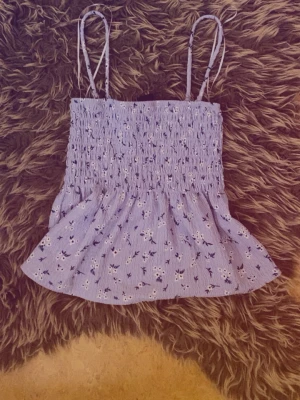 Ljusblå blommig smockad topp - Supersöt ljusblå topp med smockad överdel och tunna axelband. Toppen har ett blommigt mönster i vitt och mörkblått samt en volang nedtill för extra flowy känsla. Perfekt för sommardagar och att styla med jeansshorts eller kjol.