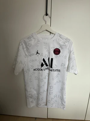 Vit PSG x Jordan matchtröja - Säljer en vit PSG x Jordan matchtröja med grått Paris-mönster över hela tröjan. Tröjan har svart tryck med Accor Live Limitless-logga och PSG-emblem i rosa och svart på bröstet. Tillverkad i lätt och ventilerande material, perfekt för fotboll eller träning. Använd ett fåtal gånger.