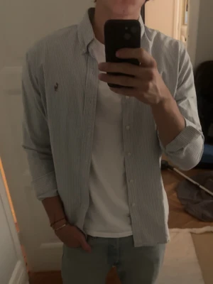 Randig skjorta från Ralph Lauren - Klassisk vit skjorta med tunna blå ränder från Ralph Lauren. Skjortan har lång ärm, knappar framtill och den ikoniska broderade loggan på bröstet. Perfekt till jeans eller chinos för en clean och stilren look. Bröstmått: 49 cm Längd: 72 cm 