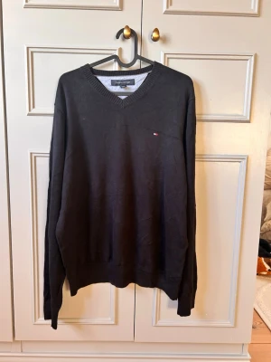 Svart stickad tröja Tommy Hilfiger - Klassisk svart stickad tröja från Tommy Hilfiger med V-ringning och diskret logga på bröstet. Tröjan har långa ärmar och ribbade muddar vid hals, ärmslut och nederkant. Perfekt till jeans eller chinos för en clean look. Helt ny oanvänd