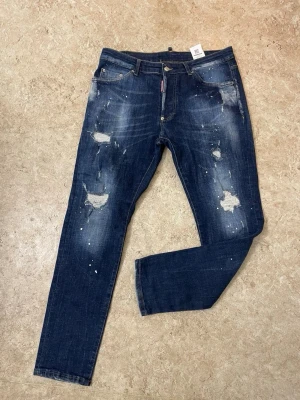 Mörkblå slitna jeans med hål - Säljer ett par mörkblå jeans med distressed detaljer och hål på benen. Jeansen har en smal passform och coola slitningar samt färgstänk för en trendig look. Perfekta för dig som gillar streetstyle och vill sticka ut.