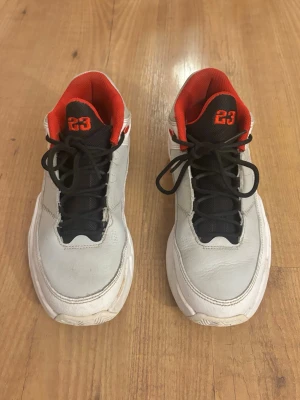 Vita Nike Jordan Max Aura 3 sneakers med röda detaljer - Säljer ett par vita Nike jordan Max aura 3 med svarta snören, svart plös och röda detaljer. Siffran 23 syns tydligt på plösen och insidan är fodrad i rött tyg. Skorna har en klassisk låg siluett och är tillverkade i läder med gummisula. Perfekt till träning eller aktiviteter. Nypris 1000