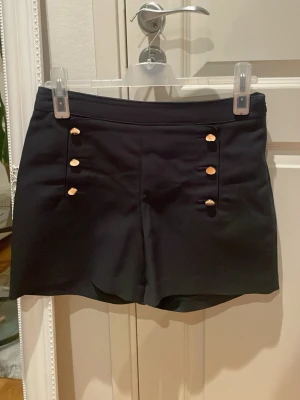 Mörka Marinblå (nästan svarta) shorts med guldknappar strl 38 - Stilrena shorts från H&M med sex snygga guldknappar framtill som ger en cool, dressad look. Klassisk passform och midwaist. Perfekta för sommaren och lätta att matcha med olika toppar.