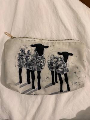 Necessär med fårmotiv - Necessär i polyester med dragkedja och tryck av två svarta får på framsidan och baksidan. Perfekt för småsaker eller smink. Unik design med konstnärlig känsla och neutrala färger som passar till det mesta. Av C.Brüggmann. Väskan har en fläck som syns på bilden vilket lär gå bort om man tvättar den, detsamma för blyertsmärkena inuti