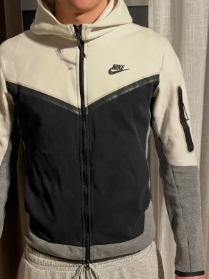 Nike hoodie med dragkedja och huva - Nike hoodie i vitt, svart och grått med dragkedja framtill och huva. Snygg V-design på bröstet, ficka med dragkedja på ärmen och Nike-logga på bröstet. Tillverkad i mjukt material, perfekt för chill eller träning.