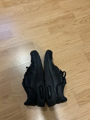Svarta Nike Air Max sneakers - Säljer ett par helsvarta Nike Air Max sneakers med klassisk låg profil och synlig Air-dämpning i sulan. Skorna har mesh och syntetmaterial på ovandelen, samt gummisula med tydligt mönster. Perfekta för dig som gillar stilrena och sportiga sneakers.