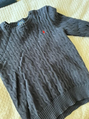 Mörkblå kabelstickad tröja Polo Ralph Lauren - Snygg mörkblå kabelstickad tröja från Polo Ralph Lauren med klassisk röd logga broderad på bröstet. Tröjan har rund halsringning, långa ärmar och breda ribbade muddar vid ärmslut och nederkant. Står L men sitter mer som xs eller S.