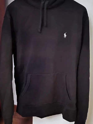 Svart hoodie från Polo Ralph Lauren - Klassisk svart hoodie från Polo Ralph Lauren med vit broderad logga på bröstet. Hoodien har huva med dragsko och en stor känguruficka framtill. Tillverkad i mjukt material som känns skönt mot huden. Perfekt för en avslappnad och stilren look.