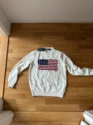 Vit stickad tröja Polo Ralph Lauren - Säljer en vit stickad tröja från Polo Ralph Lauren med amerikansk flagga broderad på bröstet och RL-detalj. Tröjan har rund halsringning och ribbade muddar vid ärmslut och nederkant. Perfekt för dig som gillar klassisk och stilren design.