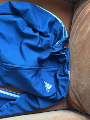 Blå Adidas ziptröja Primegreen - Snygg blå ziptröja från Adidas med vit logga på bröstet och klassiska vita ränder längs ärmarna. Tillverkad i Primegreen-material som är återvunnet. Perfekt för en sportig look och enkel att matcha med andra plagg.