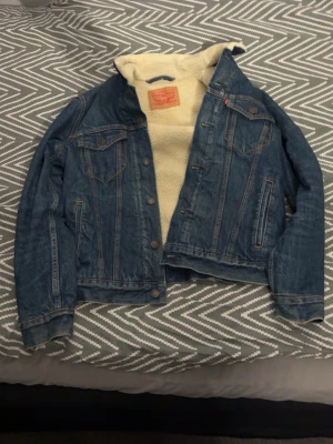 Levi's blå fodrad jeansjacka - Klassisk blå jeansjacka från Levi's med mysigt teddyfoder. Jackan har två bröstfickor med lock, sidofickor och knappar framtill. Snygga söm-detaljer och ikonisk röd Levi's-tagg vid bröstfickan. Perfekt för en cool och avslappnad stil.