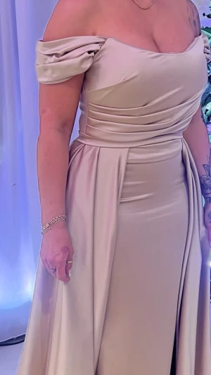 Beige långklänning med slits - Elegant beige långklänning i ett glansigt, följsamt tyg. Klänningen har en hög slits framtill och ett svepande fall som ger en lyxig känsla. Perfekt för dig som vill sticka ut på festen med en stilren och modern look.