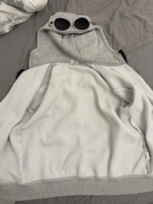 Grå hoodie med goggles från C.P. Company - Säljer en grå zip hoodie från C.P. Company med ikoniska goggles i huvan. Hoodien har två fickor fram, ribbade muddar och mjukt fleecefoder på insidan. Perfekt för dig som vill sticka ut med en unik detalj på huvan.