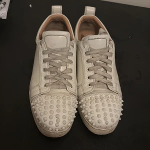 Vita Christian Louboutin sneakers med nitar - Säljer ett par vita sneakers från Christian Louboutin i skinn med ikoniska röda sulor. Skorna har snörning och coola nitar över tån för en edgy vibe. Låg modell med rund tå och klassisk siluett. Perfekta för dig som vill sticka ut med lyxig streetstyle.jätte bekväma !! Tveka inte på att höra av dig vid intresse
