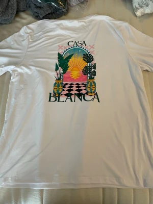 Vit Casa Blanca t-shirt med tryck - Vit t-shirt från Casa Blanca med färgglatt grafiskt tryck på ryggen och mindre tryck fram. Motivet visar en solnedgång, växter och vaser i starka färger. Klassisk passform och rund halsringning. Perfekt för dig som gillar unika prints och streetwear vibes.