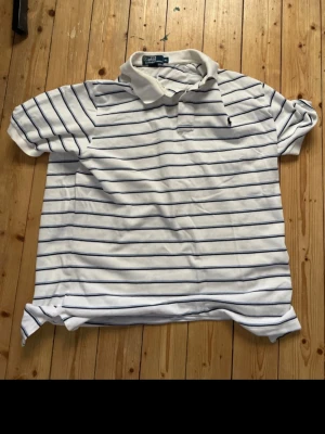 Vit randig pikétröja från Polo Ralph Lauren - Vit pikétröja med tunna blå och mörkblå ränder från Polo Ralph Lauren. Klassisk krage och knappar framtill, broderad logga på bröstet. Finns ett liten skada se bild två. 
