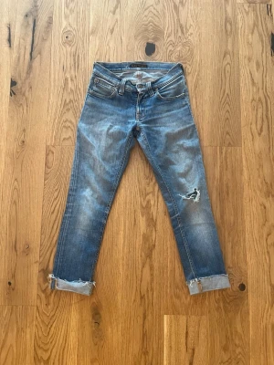 Blå slim jeans från Nudie Jeans - Säljer ett par blå jeans från Nudie Jeans med slitningar och hål på ena benet. Jeansen har fem fickor, uppvikta benslut och klassisk knappgylf. Materialet är denim i bomull och passformen är smal. Perfekta för en avslappnad och trendig look.