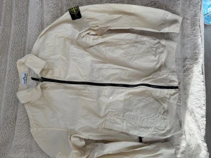 Vit vindjacka från Stone Island - Säljer en stilren vit vindjacka från Stone Island med svart dragkedja och klassisk patch på ärmen. Jackan har krage, två sidofickor och är tillverkad i ett lätt, prassligt material som passar perfekt för blåsiga dagar. Enkel och modern design som funkar till det mesta.