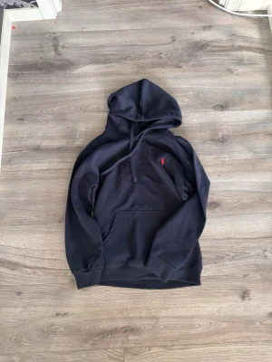 Mörkblå hoodie från Polo Ralph Lauren - Snygg mörkblå hoodie från Polo Ralph Lauren med klassisk huva och dragsko. Ikoniska röda loggan broderad på bröstet och stor magficka framtill. Perfekt för en avslappnad och stilren look.