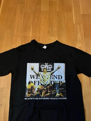 Svart Weekend Offender t-shirt - Svart t-shirt från Weekend Offender med stort grafiskt tryck på framsidan som visar en person med utsträckta armar och texten 'WE DON'T LIKE OUTSIDERS WEEKEND OFFENDER'. Rund hals och korta ärmar. Perfekt för dig som gillar streetwear och statement-plagg.