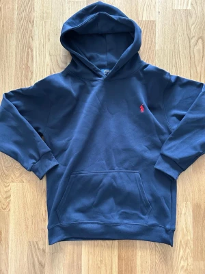 Mörkblå hoodie från Polo Ralph Lauren - Snygg mörkblå hoodie från Polo Ralph Lauren med klassisk huva och känguruficka. Ikoniska röda loggan broderad på bröstet. Tillverkad i mjuk bomullsblandning som känns skön mot huden. Perfekt för en avslappnad och stilren look.