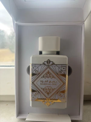 Badee Al Oud Eau de Parfum 100ml - Badee Al Oud från Lattafa är en Eau de Parfum i en elegant vit flaska med gulddetaljer och geometriskt mönster. Flaskan rymmer 100 ml och har en modern, fyrkantig form med matchande vit kork. Doften är orientalisk och exklusiv, perfekt för dig som gillar unika parfymer.