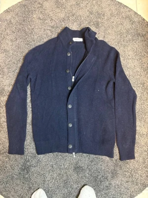 Mörkblå cardigan Gran Sasso - Mörkblå kofta från Gran Sasso med ståkrage och knäppning framtill. Koftan har långa ärmar, ribbade muddar och är tillverkad i ett mjukt stickat material. Perfekt för lager-på-lager och enkel att matcha med olika outfits.