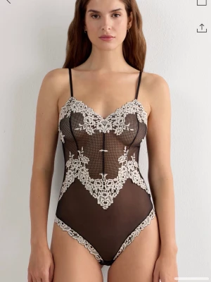 Svart spetsbody med vita broderier - Snygg svart mesh-body med tunna axelband och detaljerade vita spetsbroderier. Djup urringning och hög benskärning ger en feminin siluett. Perfekt för dig som gillar eleganta underkläder med en lyxig känsla.