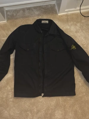 Svart overshirt från Stone Island - Snygg marin blå overshirt från Stone Island med klassisk patch på vänster ärm. Två bröstfickor med dragkedja. Tillverkad i slitstarkt bomullsmaterial, perfekt för lager på lager och streetwear-stil. Jätte bra skick och inga skador passar till vad som helst. Skriv för fler bilder och andra frågor 