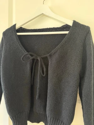 Mörkgrå stickad cardigan med knyt - Mysig mörkgrå stickad cardigan med långa ärmar och knytband framtill. Tröjan har en djup v-ringning och ribbstickade muddar vid ärmslut och nederkant. Perfekt att slänga över en topp för en avslappnad look.