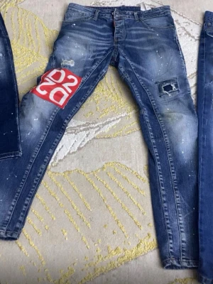 DSQUARED2 blå skinny jeans - Säljer ett par blå skinny jeans från DSQUARED2 med slitningar, patchdetaljer och vit färgstänk. Ena benet har en stor röd och vit DSQ2-logga. Jeansen har klassisk femficksmodell och dragkedjegylf. Perfekta för dig som vill sticka ut!