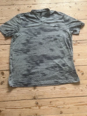 Grå sportpiké från Adidas - Snygg grå sportpiké från Adidas med korta ärmar och diskret mönster i olika grå nyanser. Pikén har två knappar vid halsen och en liten Adidas-logga på bröstet. Perfekt för träning eller chill dagar.