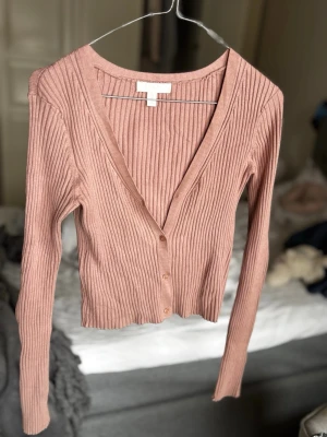 Rosa ribbad kofta från H&M - Snygg rosa ribbad kofta från H&M med djup V-ringning och knappar framtill. Modellen är croppad med långa ärmar och smal passform, perfekt att styla över ett linne eller t-shirt. Materialet är mjukt och stretchigt för extra komfort.