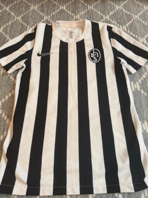 Randig fotbollströja från Nike - Snygg svartvit randig fotbollströja från Nike med klubbmärke på bröstet och klassisk rund hals. Perfekt för träning eller match. Tillverkad i lätt och ventilerande material som passar bra för sportaktiviteter.