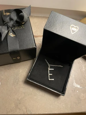 Maria Nilsdotter halsband - Silver halsband från Maria Nilsdotter med ett E. Nypris ca 2000. Del av kedjan har tyvärr gått sönder så man behöver sätta i kedjan från båda hållen i cirkeln. Men det är inget som blir problem. Kedjan är 45 cm. Förpackningen kommer med. 