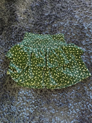 Grön blommig volangkjol - Säljer en grön kortkjol med vita blommor och volangdetaljer. Kjolen har bred smock i midjan som ger en bekväm passform och ett snyggt fall. Perfekt för dig som gillar en lekfull och trendig stil.