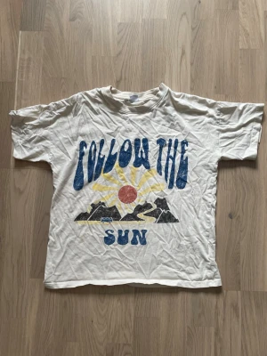 Vit t-shirt med tryck från Lindex - Vit t-shirt från Lindex med stort färgglatt tryck på framsidan där det står 'Follow the Sun' och motiv av sol, berg och väg. Klassisk rund hals och korta ärmar. Tillverkad i mjuk bomull, perfekt för en avslappnad stil.