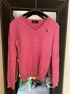 Rosa kabelstickad tröja Polo Ralph Lauren - Kabelstickad rosa tröja från Polo Ralph Lauren med V-ringning och den klassiska broderade loggan i marinblått på bröstet. Tröjan har långa ärmar och ribbade muddar vid ärmslut och nederkant. Perfekt för dig som gillar en preppy och färgglad stil.