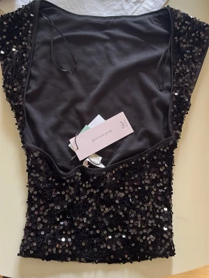 Svart paljettopp från Gina Tricot - Supersnygg svart topp från Gina Tricot täckt med glittrande paljetter. Modellen är ärmlös med rund halsringning och har en figurnära passform. Perfekt för dig som vill sticka ut och glänsa lite extra. Materialet är mjukt och stretchigt för skön känsla.