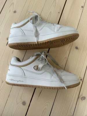 Vita Champion sneakers med beige detaljer - Snygga vita sneakers från Champion med beige detaljer och gummisula. Skorna har klassisk låg profil, vita skosnören och meshpartier för extra andningsförmåga. Champion-logga på sidan och hälen ger en sportig vibe. Perfekta för dig som gillar stilrena och bekväma sneakers. I storlek 37,5 (23,5cm). Några smutsfläckar, se bilderna, annars inga slitningar eller liknande defekter