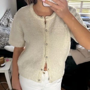 Krämvit kortärmad kofta med pärlknappar - Söt krämvit kofta med korta ärmar och rund halsringning. Koftan har dekorativa pärlknappar framtill och är stickad i ett mjukt, fluffigt material som ger en mysig känsla. Perfekt att styla med jeans eller kjol för en trendig look.