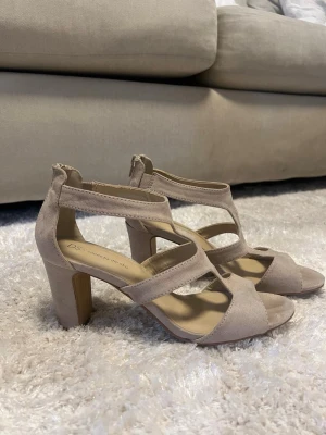 Beige sandaler med klack och remmar - Säljer ett par snygga beige sandaler med bred klack och flera remmar över foten. Sandalerna har öppen tå och dragkedja bak för enkel påtagning. Tillverkade i mockaimitation som ger en mjuk och stilren look.