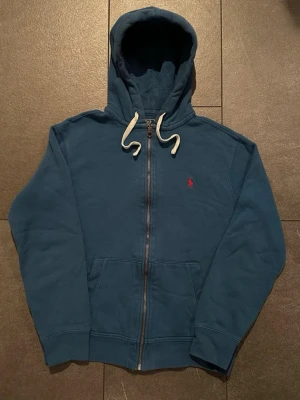 Blå zip hoodie från Polo Ralph Lauren - Mörkblå hoodie med dragkedja från Polo Ralph Lauren. Klassisk huva med vita snören och röd broderad logga på bröstet. Mjuk bomullskvalitet och kängurufickor framtill. Perfekt för en avslappnad och stilren look.