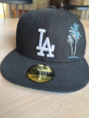 Svart LA Dodgers keps från New Era - Säljer en svart 59FIFTY keps från New Era med LA Dodgers-logga i vitt och broderade palmer i blått och vitt framtill. På sidan finns ett jubileumsmärke och en broderad arena. Klassisk flat brim och snapback-stil.